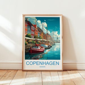 Op de afbeelding: Een ingelijste kunstdruk met een levendige illustratie van Kopenhagen, Denemarken. Het kunstwerk toont kleurrijke gebouwen langs een kanaal met verschillende zeilboten. Het woord "KOPENHAGEN" is in grote blauwe letters gedrukt, met "DENEMARKEN" eronder.