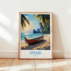 May include: A framed art print of a tropical beach scene. A wooden boat rests on the sandy shore, with palm trees and cliffs in the background. The text "KRABI THAILAND" is printed at the bottom of the image. The artwork is in a wooden frame.