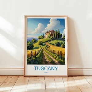 May include: Framed art print of a Tuscan landscape. The artwork depicts a winding dirt road leading to a villa on a hilltop, surrounded by green trees and fields of golden wheat under a blue sky. The word "TUSCANY" is printed at the bottom.