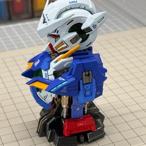 May include: A detailed model of a robot's head and upper torso, featuring a white helmet with blue and red accents. The robot has blue and gold armor, with a red component at the base. The model is on a gray surface.