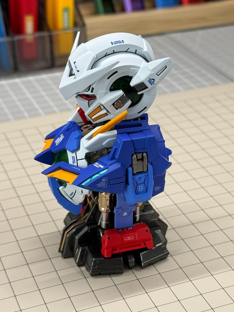 May include: A detailed model of a robot's head and upper torso, featuring a white helmet with blue and red accents. The robot has blue and gold armor, with a red component at the base. The model is on a gray surface.
