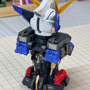 May include: A partially assembled Gundam model kit. The model features a white head with blue and yellow accents, black shoulders, blue arms, and red feet. The model is set against a grid background.