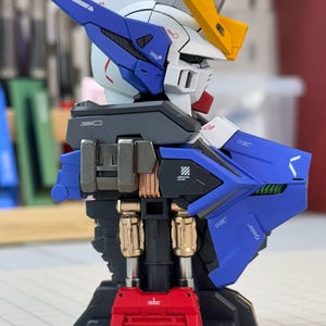May include: A detailed model of a robot's upper body, featuring a white head with blue and yellow accents. The robot has a blue chest and back, with metallic gold and silver components. A red base supports the structure, showcasing intricate mechanical details.