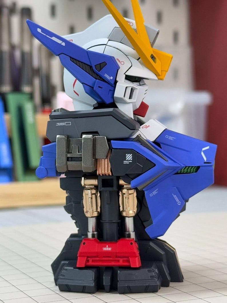 May include: A detailed model of a robot's upper body, featuring a white head with blue and yellow accents. The robot has a blue chest and back, with metallic gold and silver components. A red base supports the structure, showcasing intricate mechanical details.