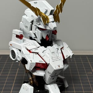 Puede incluir: Modelo detallado en blanco y dorado de un busto de robot. El modelo presenta paneles intrincados, detalles en rojo y cuernos dorados. El busto se exhibe sobre una superficie oscura.