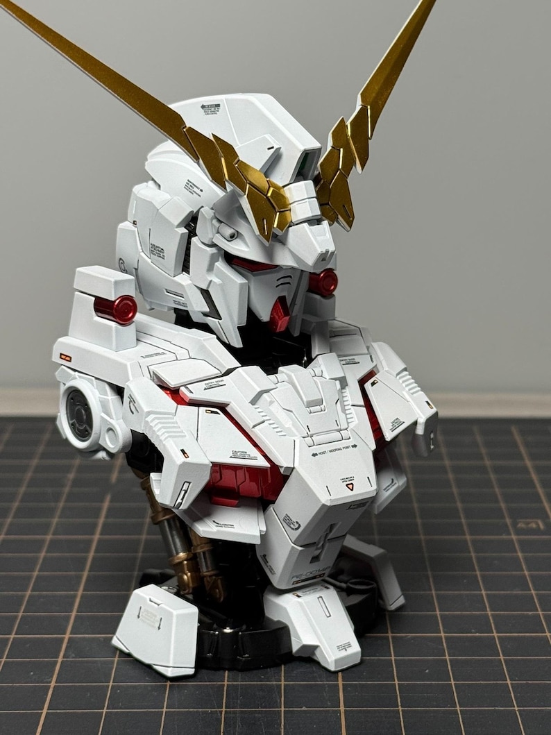 Puede incluir: Modelo detallado en blanco y dorado de un busto de robot. El modelo presenta paneles intrincados, detalles en rojo y cuernos dorados. El busto se exhibe sobre una superficie oscura.