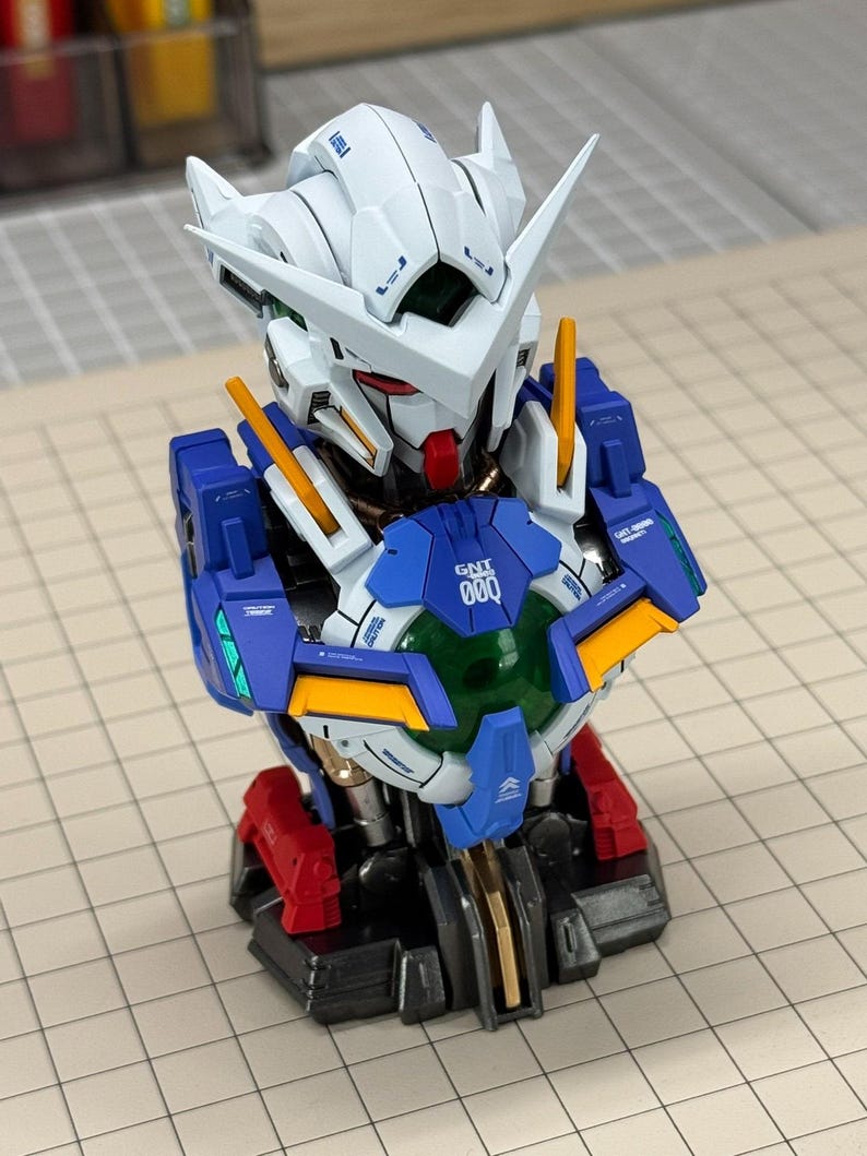 May include: A detailed model of a Gundam robot bust. The bust features a white head with sharp angles, blue and red accents, and the text "GNT 000" on the chest. The model is on a grid-lined surface.