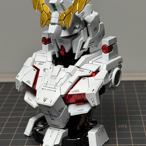 Puede incluir: Modelo detallado de un busto de robot Gundam. El modelo es principalmente blanco con detalles en rojo y dorado. La cabeza presenta detalles intrincados y una prominente cresta dorada. El busto se exhibe sobre una base negra.