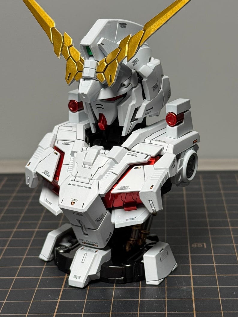 Puede incluir: Modelo detallado de un busto de robot Gundam. El modelo es principalmente blanco con detalles en rojo y dorado. La cabeza presenta detalles intrincados y una prominente cresta dorada. El busto se exhibe sobre una base negra.