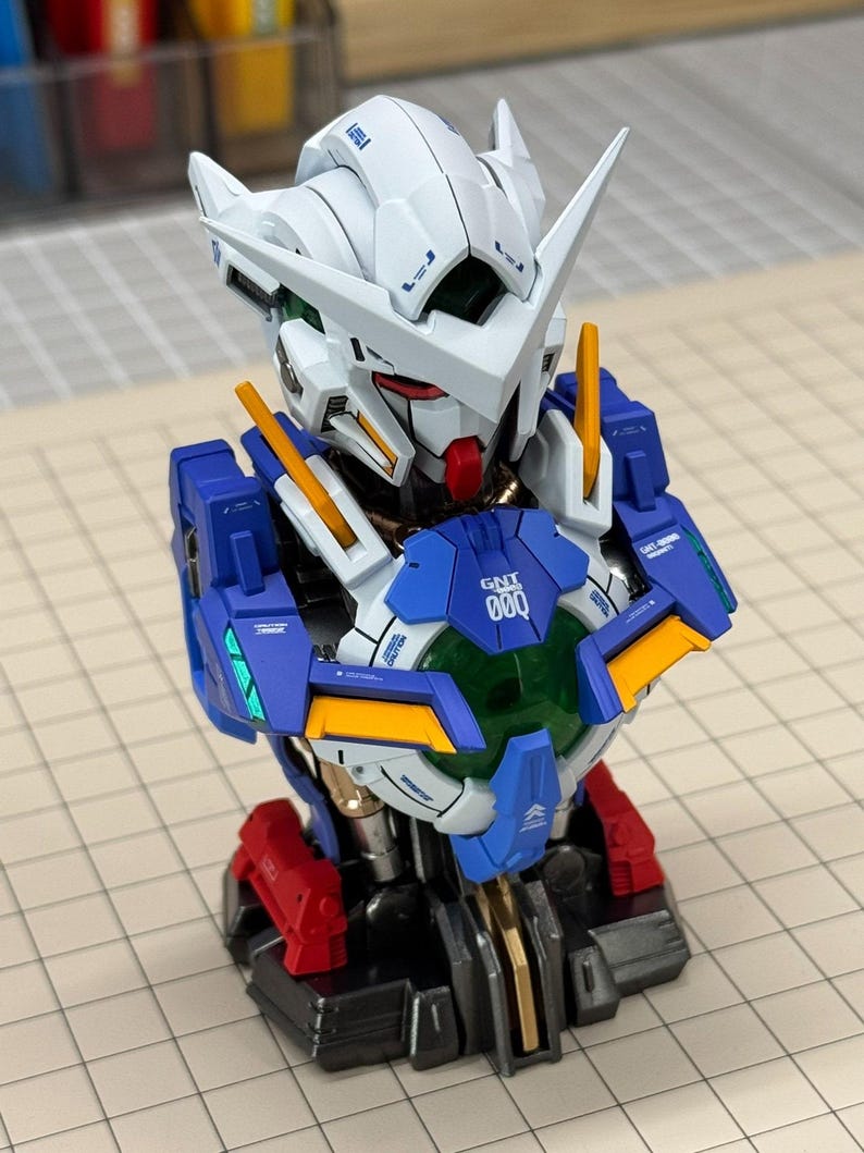 May include: A detailed model of a Gundam robot bust, featuring a white head with blue and red accents. The robot has a distinctive design with sharp angles and intricate details, including yellow and orange highlights. The chest area displays the text "GNT 000".