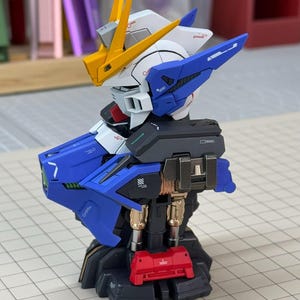May include: A detailed model of a Gundam robot, featuring a color scheme of blue, white, yellow, and red. The model has intricate mechanical details, including articulated joints and panels. The robot is posed on a grid surface, showcasing its design.