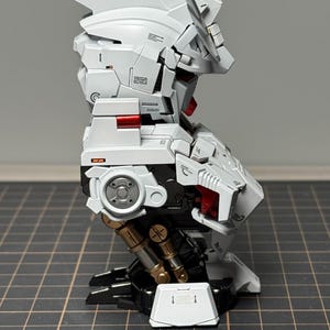 Puede incluir: Un kit de modelo blanco y negro parcialmente ensamblado con detalles en rojo y dorado. El modelo presenta intrincados detalles mec&aacute;nicos, incluyendo articulaciones y paneles articulados. El kit se encuentra sobre un fondo de cuadr&iacute;cula, mostrando su complejo dise&ntilde;o.