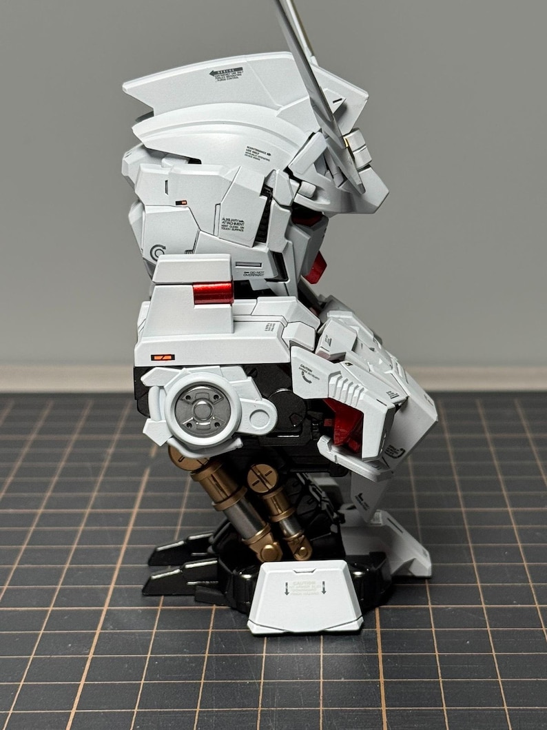 Puede incluir: Un kit de modelo blanco y negro parcialmente ensamblado con detalles en rojo y dorado. El modelo presenta intrincados detalles mec&aacute;nicos, incluyendo articulaciones y paneles articulados. El kit se encuentra sobre un fondo de cuadr&iacute;cula, mostrando su complejo dise&ntilde;o.