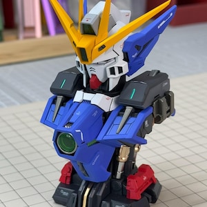 May include: A detailed model of a robot with a blue, white, yellow, black, and red color scheme. The robot has a complex design with multiple panels, sharp angles, and a prominent head with yellow antennae. The model is on a grid surface.