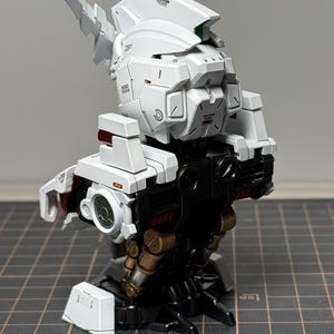 Puede incluir: Maqueta de robot parcialmente ensamblada, en blanco y negro. El modelo presenta detalles intrincados, incluyendo paneles, articulaciones y componentes mec&aacute;nicos. Acentos rojos y dorados a&ntilde;aden contraste. El modelo est&aacute; sobre un fondo neutro.