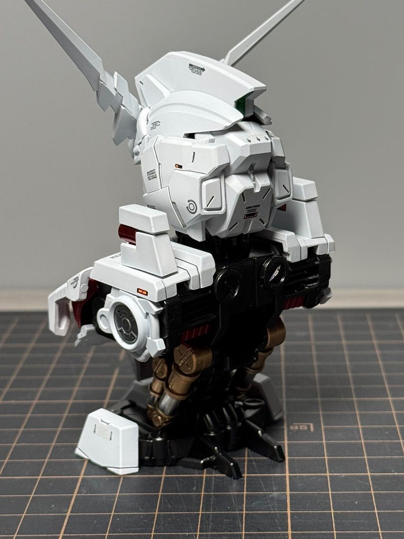 Puede incluir: Maqueta de robot parcialmente ensamblada, en blanco y negro. El modelo presenta detalles intrincados, incluyendo paneles, articulaciones y componentes mec&aacute;nicos. Acentos rojos y dorados a&ntilde;aden contraste. El modelo est&aacute; sobre un fondo neutro.