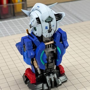 May include: A partially assembled model robot with a white head, blue torso, and red accents. The robot has intricate details and mechanical components, suggesting a hobbyist project or collectible item. The model is set against a grid background.