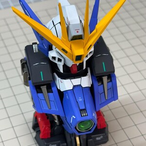May include: A detailed model of a Gundam robot, featuring a blue, white, yellow, red, and black color scheme. The model has intricate details, including angular armor, a head with distinctive yellow fins, and a green lens. It is a collectible item.