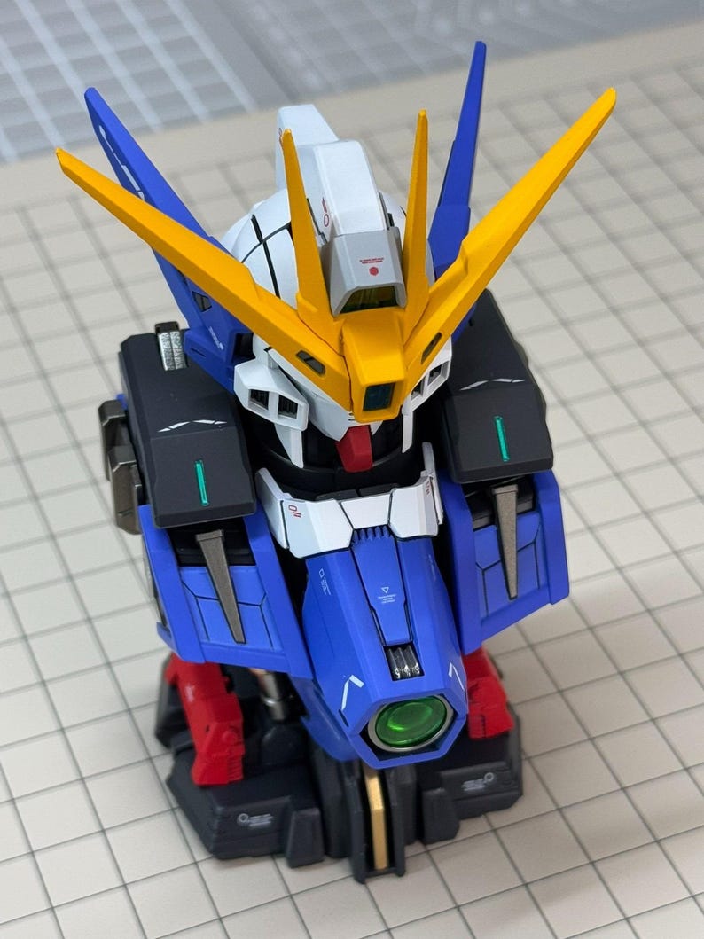 May include: A detailed model of a Gundam robot, featuring a blue, white, yellow, red, and black color scheme. The model has intricate details, including angular armor, a head with distinctive yellow fins, and a green lens. It is a collectible item.