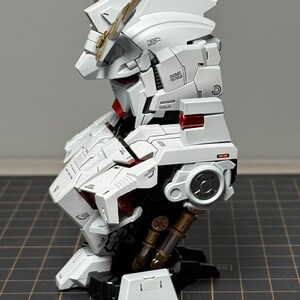 Puede incluir: Modelo detallado de robot blanco y dorado, con intrincados paneles y detalles en rojo. El modelo tiene una cabeza estilizada con cuernos dorados y est&aacute; montado sobre una base negra. El dise&ntilde;o muestra una est&eacute;tica futurista.