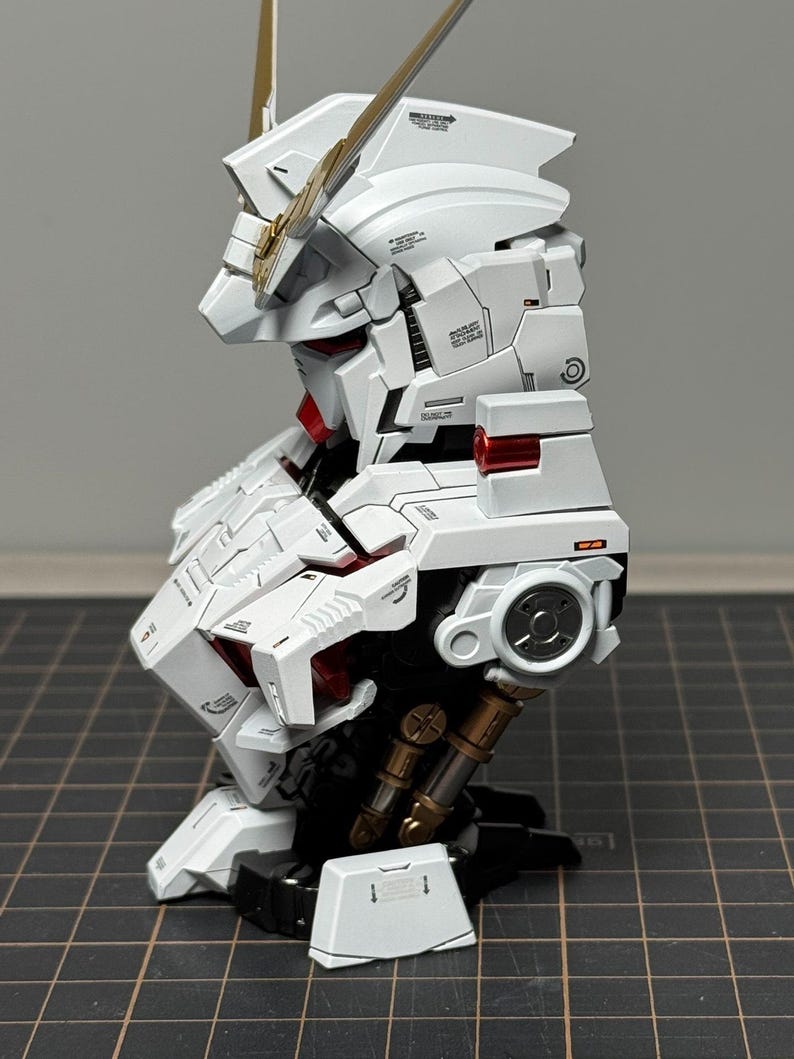 Puede incluir: Modelo detallado de robot blanco y dorado, con intrincados paneles y detalles en rojo. El modelo tiene una cabeza estilizada con cuernos dorados y est&aacute; montado sobre una base negra. El dise&ntilde;o muestra una est&eacute;tica futurista.