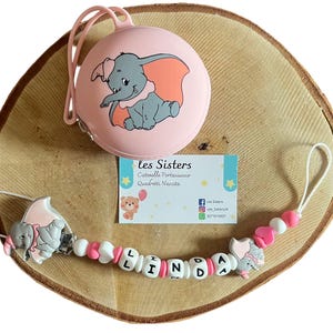 Peut inclure: Ensemble de porte-sucette et attache-tétine roses avec un motif Dumbo l'éléphant. L'attache-tétine épelle le nom "LINDA" avec des perles de lettres blanches et des accents roses. L'ensemble est présenté sur une surface en bois.
