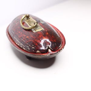 Pode incluir: Uma caixa de joias oval em laca vermelha escura com acabamento brilhante e textura manchada. A tampa apresenta uma alça de metal cor de latão fixada a uma placa retangular. A caixa tem uma pequena abertura em entalhe numa extremidade.