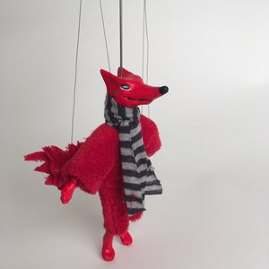 May include: Red fox puppet with a black and white striped scarf. The puppet has a red fuzzy body and a red painted face.