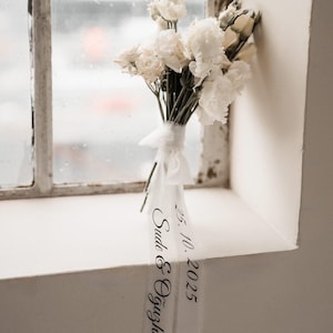 Op de afbeelding: Een boeket witte bloemen, vastgebonden met een wit lint, rust op een vensterbank. Het lint heeft zwarte tekst met de tekst "25. 10. 2025 Sude & Oğuzhan". Het raam is wit met een bewolkte achtergrond.