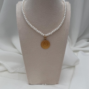 Pode incluir: Um colar de pérolas com um pendente de ouro exposto em um manequim. O colar apresenta pequenas pérolas redondas e um pendente circular de ouro com um design detalhado. O fundo é um tecido branco macio.