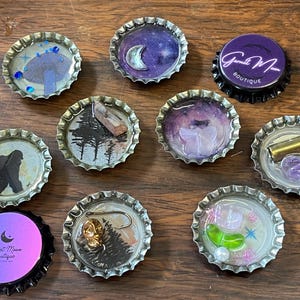 Peut inclure: Une collection de bouchons de bouteilles décoratifs avec divers motifs. Certains bouchons présentent des images de champignons, de croissant de lune, de forêt et de silhouette de Bigfoot. D'autres ont des objets intégrés comme des cristaux et un leurre de pêche. Un bouchon porte le texte "Garnet Moon Boutique".