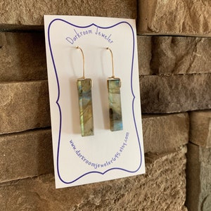 以下が含まれることがあります： 虹色のラブラドライトの四角い石がついた、ゴールドのドロップイヤリングのペア。イヤリングは、"Darkroom Jeweler"というテキストとウェブサイトのアドレスが書かれた白いカードに飾られています。