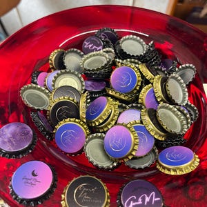 May include: A collection of bottle caps with a celestial design, featuring a crescent moon and the text "Garnet Moon Boutique." The caps have a metallic gold edge and a gradient of purple, pink, and blue colors. They are displayed in a red glass bowl.