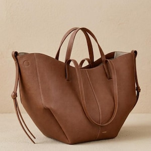 Puede incluir: Un bolso tote de cuero marr&oacute;n con dos asas cortas y dos correas para el hombro m&aacute;s largas. El bolso tiene un dise&ntilde;o estructurado con costuras visibles y un logotipo sutil cerca de la base. El bolso es de tama&ntilde;o mediano y parece estar vac&iacute;o.