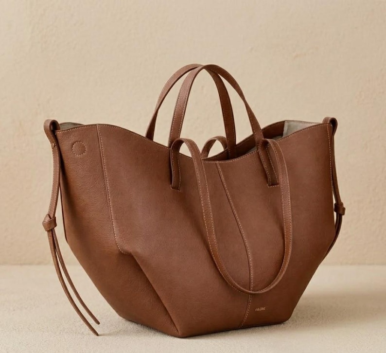 Puede incluir: Un bolso tote de cuero marr&oacute;n con dos asas cortas y dos correas para el hombro m&aacute;s largas. El bolso tiene un dise&ntilde;o estructurado con costuras visibles y un logotipo sutil cerca de la base. El bolso es de tama&ntilde;o mediano y parece estar vac&iacute;o.