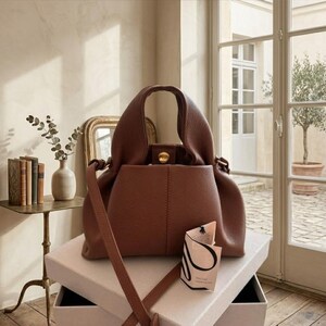 Könnte beinhalten: Eine braune Lederhandtasche mit einem oberen Griff und einem Schulterriemen, die auf einer weißen Box liegt. Die Tasche hat ein strukturiertes Design mit einem goldfarbenen Verschluss. Eine kleine Parfümbox befindet sich vor der Tasche. Links ein kleiner Beistelltisch mit Büchern und einer Vase mit Pflanzen.