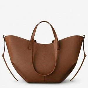 Puede incluir: Un bolso tote de cuero marr&oacute;n con dos asas y correas ajustables para los hombros. El bolso tiene una abertura ancha y un dise&ntilde;o estructurado. El bolso est&aacute; hecho de un material de cuero liso y mate. El bolso es de color neutro.