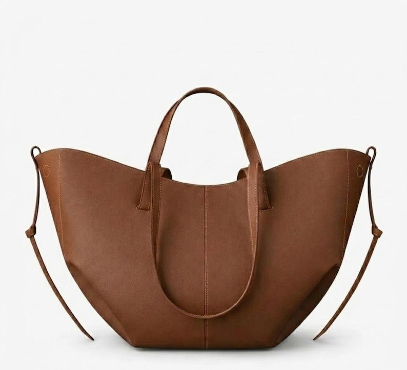 Puede incluir: Un bolso tote de cuero marr&oacute;n con dos asas y correas ajustables para los hombros. El bolso tiene una abertura ancha y un dise&ntilde;o estructurado. El bolso est&aacute; hecho de un material de cuero liso y mate. El bolso es de color neutro.