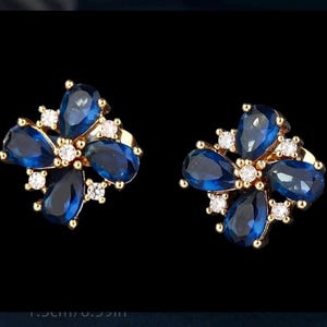 May include: A pair of gold-toned flower-shaped earrings. Each earring features five teardrop-shaped, deep blue gemstones and small, round, clear gemstones. The earrings are approximately 0.59 inches in diameter.