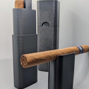 May include: A black cigar holder and case set. The set includes a holder with a cigar, a case, and a separate cigar rest. The cigar is brown with a blue band. The items are on a light gray surface.
