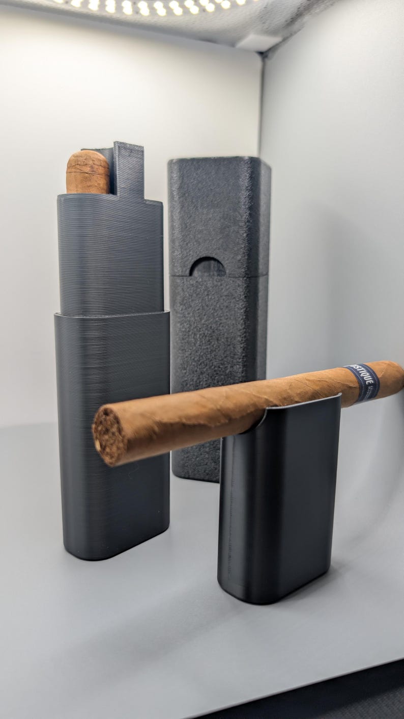 May include: A black cigar holder and case set. The set includes a holder with a cigar, a case, and a separate cigar rest. The cigar is brown with a blue band. The items are on a light gray surface.