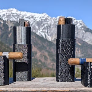 May include: Cigar holders and cases in black with textured designs. The cases are cylindrical and rectangular, with cigars resting in the holders. The background features a mountain range under a clear blue sky.