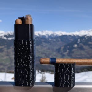 May include: A black cigar case and stand with a textured pattern. The case holds two cigars and a lighter. A cigar with a blue band rests on the stand. The background features a snowy mountain range.