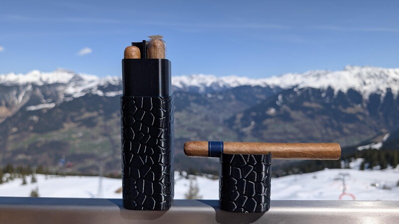 May include: A black cigar case and stand with a textured pattern. The case holds two cigars and a lighter. A cigar with a blue band rests on the stand. The background features a snowy mountain range.