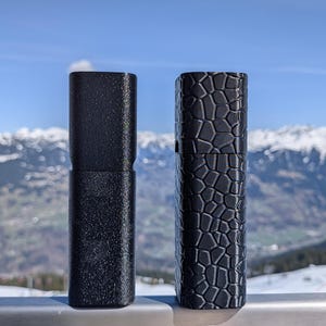 May include: Two black cylindrical cases stand side-by-side against a backdrop of snow-capped mountains and a blue sky. One case has a textured surface, while the other features a geometric pattern. The cases appear to be made of a durable material.