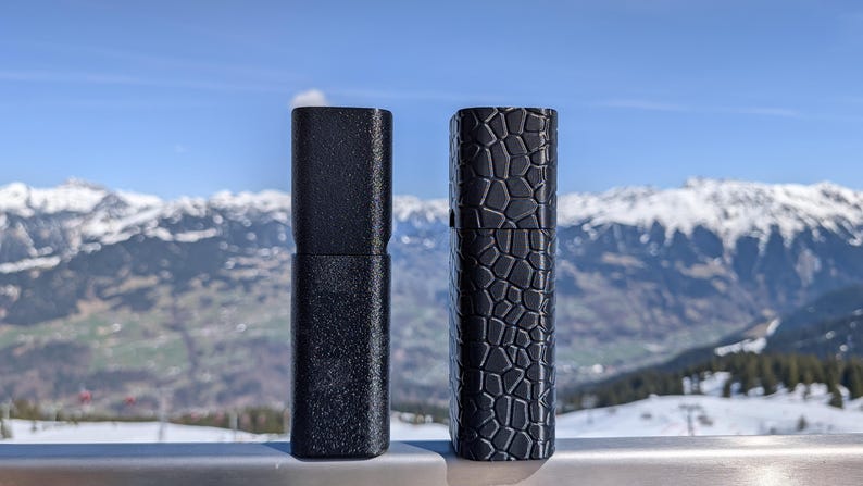 May include: Two black cylindrical cases stand side-by-side against a backdrop of snow-capped mountains and a blue sky. One case has a textured surface, while the other features a geometric pattern. The cases appear to be made of a durable material.