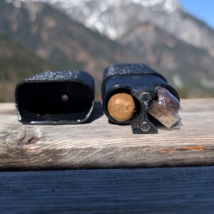 May include: Two black, textured, rectangular containers sit on a wooden surface. One is open, revealing a brown sphere and a wrapped item. The other is empty. The background features a mountain range under a blue sky.