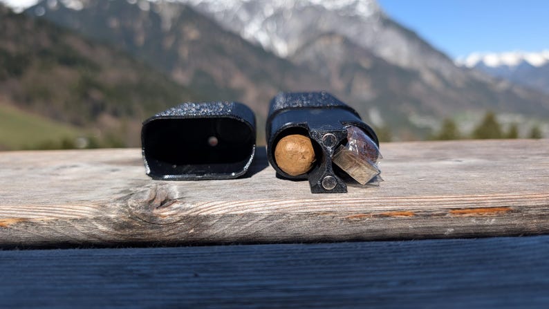 May include: Two black, textured, rectangular containers sit on a wooden surface. One is open, revealing a brown sphere and a wrapped item. The other is empty. The background features a mountain range under a blue sky.