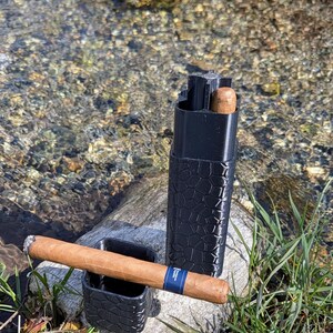 May include: A black, textured cigar case and ashtray set with a lit cigar resting in the ashtray. The cigar has a blue band. A second cigar is visible inside the case. The items are on a rock near a stream.