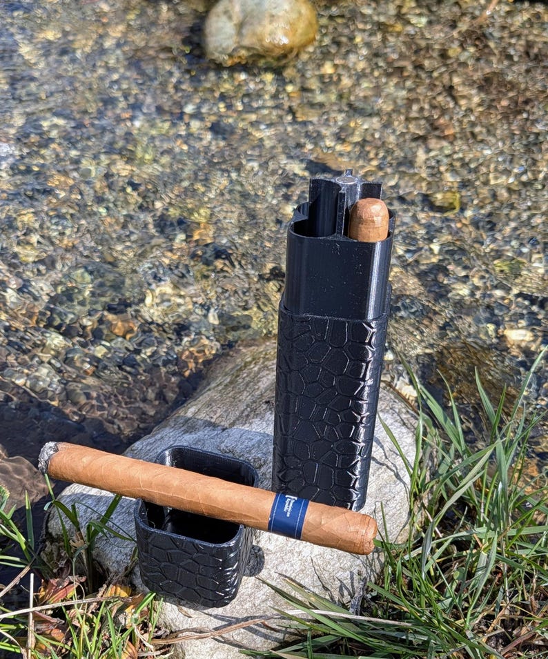 May include: A black, textured cigar case and ashtray set with a lit cigar resting in the ashtray. The cigar has a blue band. A second cigar is visible inside the case. The items are on a rock near a stream.